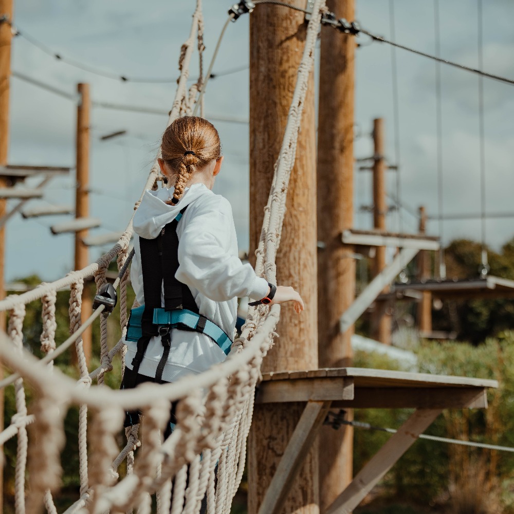 Girl in safety harness crossing a rope bridge on the High Ropes Aerial Adventure Course at Aztec Adventure The Lost Valley
