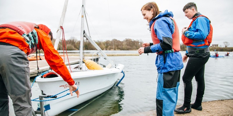 Instructor launching a dinghy from the slipway as two young sailors in buoyancy aids look on during an RYA sailing course at Upton Warren