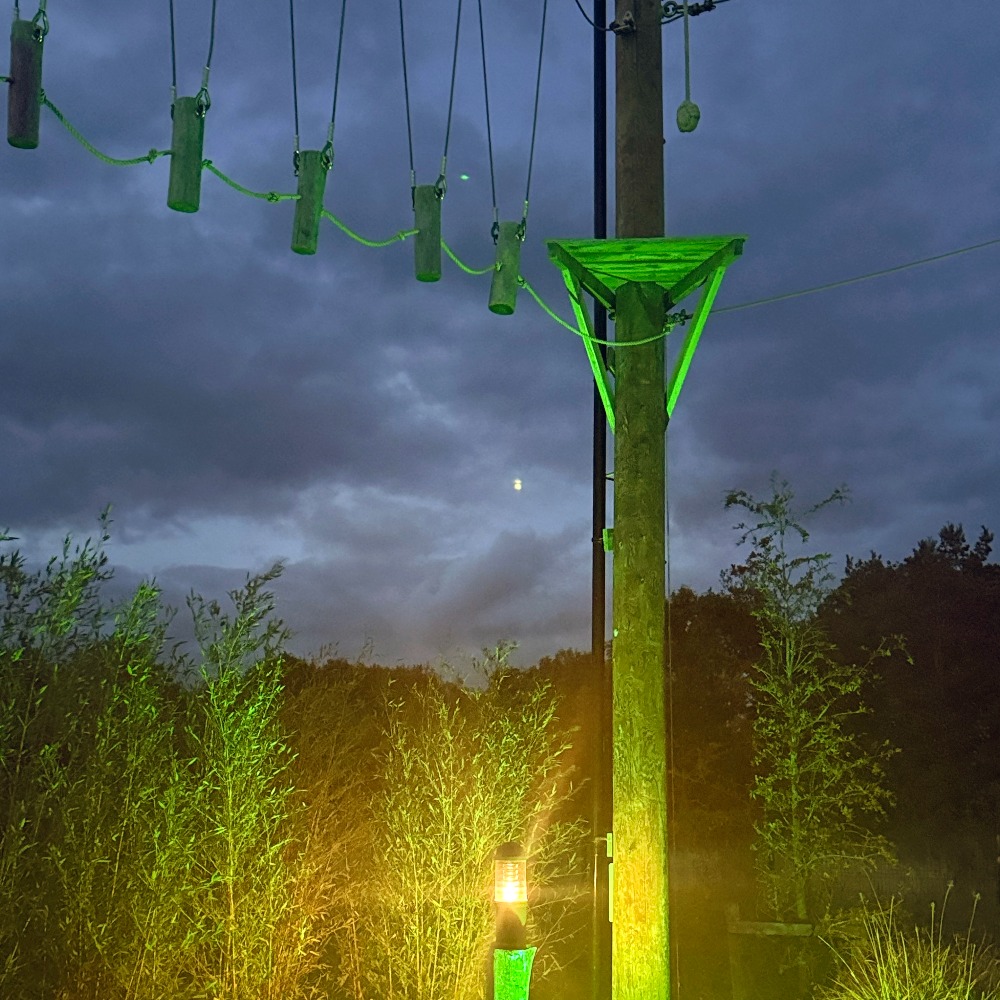 Neon Heights high ropes aerial adventure course glowing with neon lights, featuring zip lines, wobbly bridges, swingy steps, and a thrilling 70-metre zip wire descent under the night sky.
