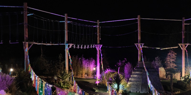 Neon Heights high ropes aerial adventure course glowing with neon lights, featuring zip lines, wobbly bridges, swingy steps, and a thrilling 70-metre zip wire descent under the night sky.