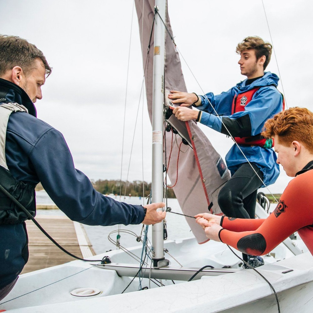 Two teenagers hoisting the sail on a dinghy with help from an instructor on a kids' RYA sailing course at Aztec Adventure Upton Warren