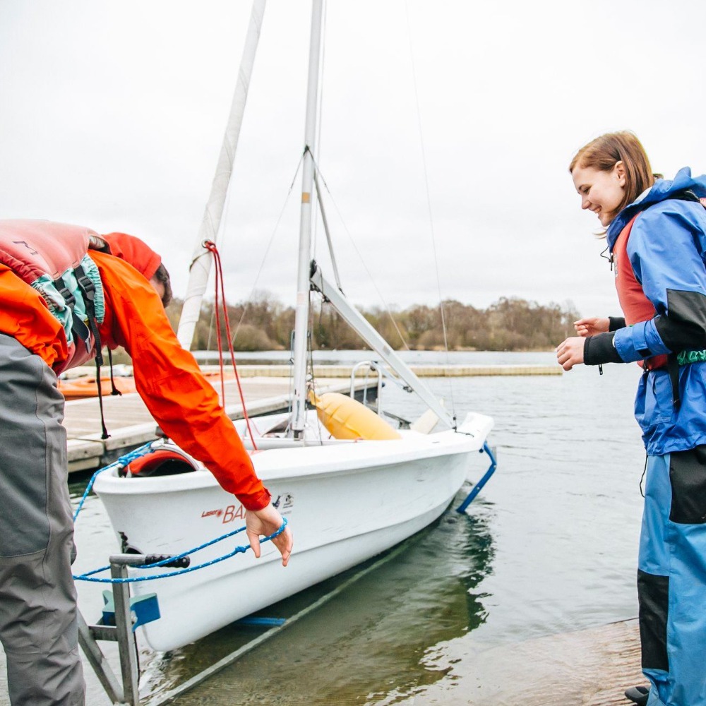 Instructor launching a dinghy from the slipway as a young sailor in buoyancy aid look on during an RYA sailing course at Upton Warren