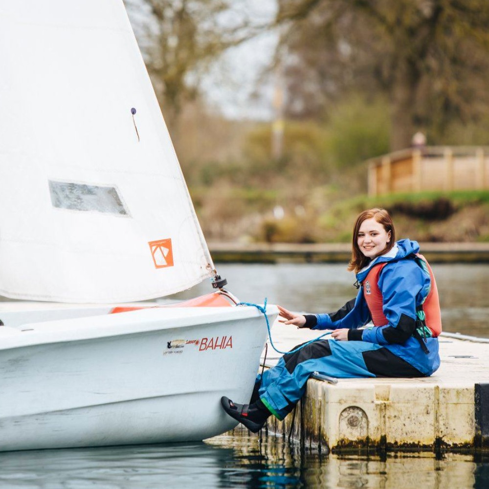 Smiling young sailor in waterproofs and a buoyancy aid sitting on the pontoon holding a dinghy's line at Upton Warren sailing lake
