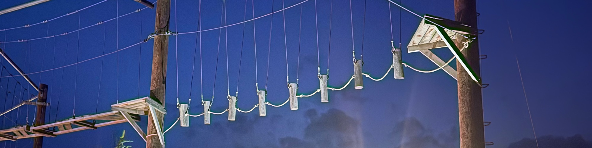 Neon Heights high ropes aerial adventure course glowing with neon lights, featuring zip lines, wobbly bridges, swingy steps, and a thrilling 70-metre zip wire descent under the night sky.